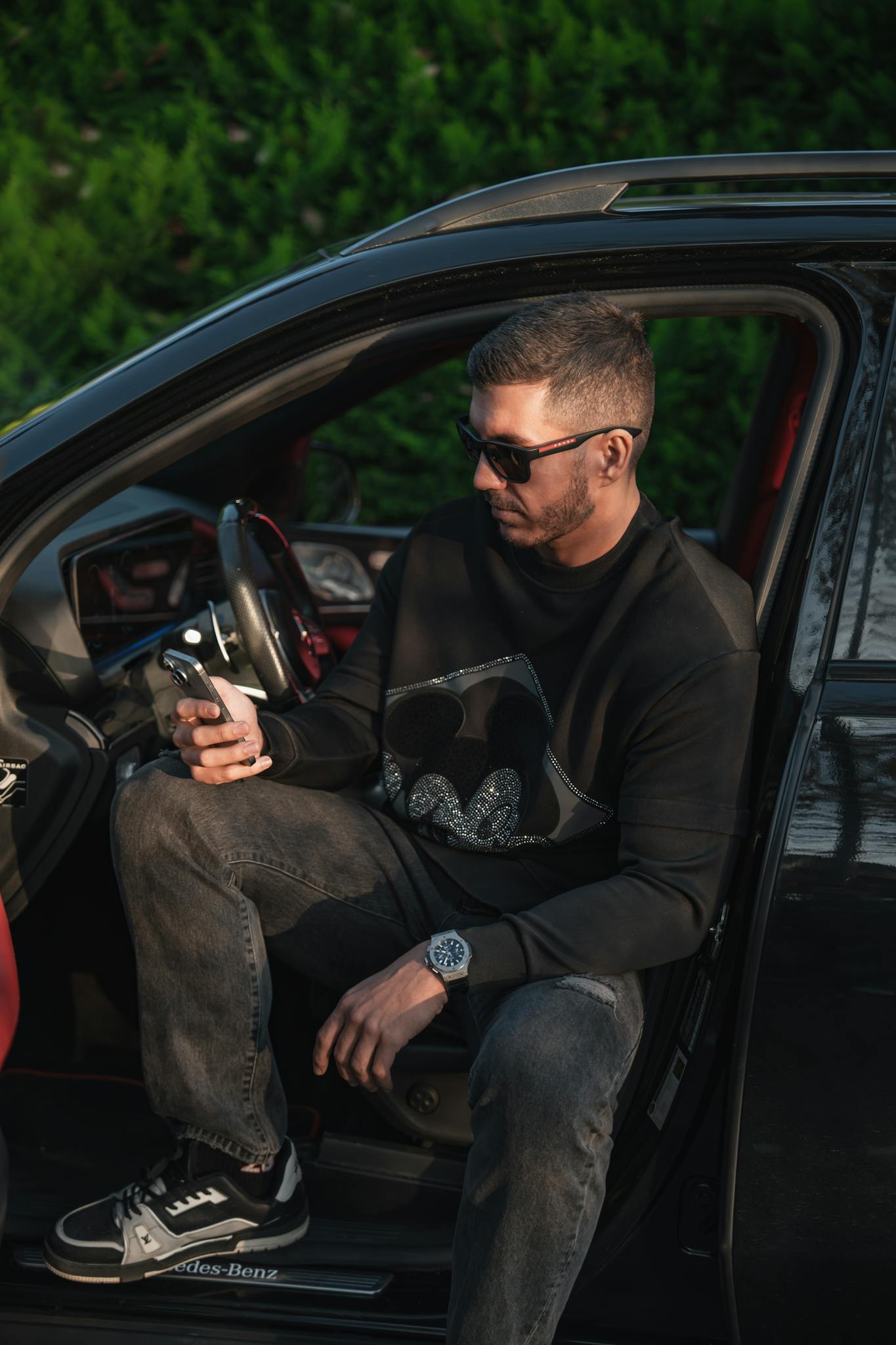Fashionable man wearing sunglasses sits in car using smartphone, exuding modern style.